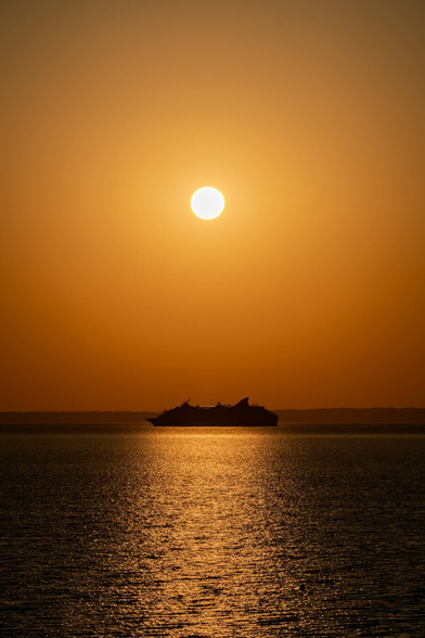 The image captures a serene sunrise over the ocean, with the sun positioned near the center of the frame, casting an orange hue across the sky. Below the sun, a dark silhouette of a large cruise ship is visible, moving across the horizon from left to right. The sunlight reflects off the surface of the water, creating a golden path that shimmers towards the foreground. The gradation of colors in the sky ranges from deep orange near the horizon to a lighter orange closer to the top of the image, indicating the rising of the sun against a clear sky.