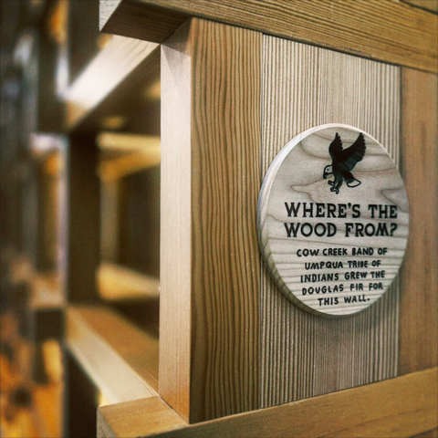 Perpendicular wooden support beams intersect off into the distance, as etched wording in a circular wooden sign in the foreground reads, "WHERE'S THE WOOD FROM? | COW CREEK BAND OF UMPQUA TRIBE OF INDIANS GREW THE DOUGLAS FIR FOR THIS WALL."