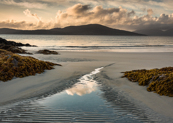 This photograph portrays a serene scene at Seilebost beach on the Isle of Harris, Outer Hebrides, Scotland, UK. The sun sets, casting a warm glow over a rivulet running through sand ripples, reflecting clouds in the sky. A distant mountain adds a touch of mystery to the tranquil setting.