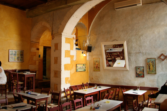 Interior of a rustic cafe with arched doorways, wooden tables and chairs, warm yellow walls, and several vintage framed pictures.