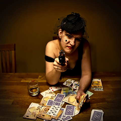 Eine schöne Frau in einem schwarzen Kostüm sitzt am Tisch, vor ihr ein Pokerblatt, viele Karten und jede Menge Geld. Einige Scheine hält sie in der linken Hand. Mit der rechten hält sie eine Pistole auf ihren Gegenüber gerichtet und lächelt siegesgewiss. 