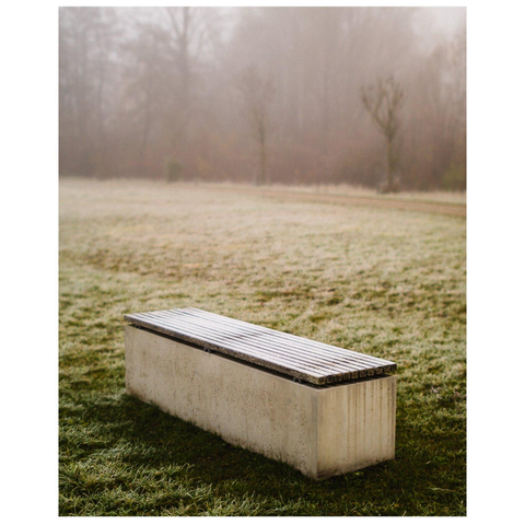 Eine Bank aus Beton steht in einem Feld. Dahinter Bäume im Nebel.