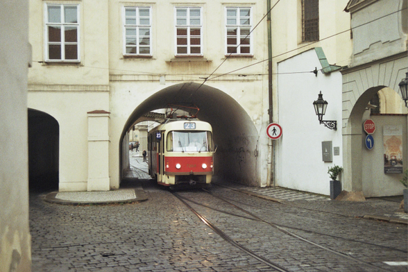 A photograph of an old style red and cream tram in Prague travelling along a brick lined street passing under an arch of a building along two tracks that almost merge together as there is only room for single file traffic.
