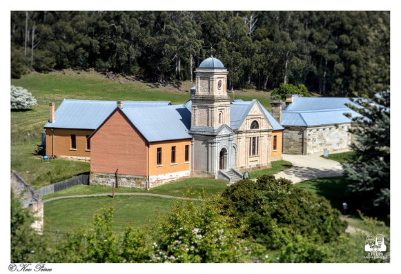 A view of the penitentiary grounds showing a prominent, two story stone building with a central clock tower and dome roof at Port Arthur historic settlement in Tasmania.

This central structure is flanked by simpler, single story buildings with grey roofs; one brick orange on the left and one rough cut stone on the right.

The buildings are set in a grassy landscape with dense green forest rising on the hill behind them.