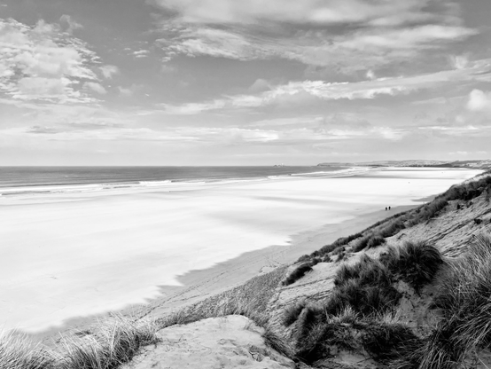 A black and white photograph taken from the top of a sand dune covered in long grass looking down to a large expanse of sand stretching to the far right hand side of the image. Out on a relatively calm sea there is a small island with a white lighthouse.