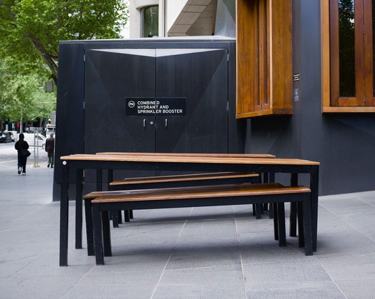 Photo of two long, outdoor, wood-and-iron cafe tables and their accompanying benches. The tables are placed outside the building where the footpath is sloping downwards towards the street. The legs on one long side of the table are taller than the legs on the other side, resulting in the eating surfaces of the tables being perfectly horizontal.