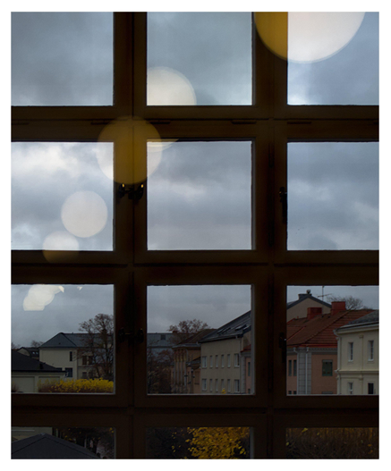 En stående bild på ett fönster från insidan. Fönstret är delat i rutor och på utsidan syns byggnader i en stadsmiljö och träd med gula löv. I glaset speglas en runda lampor i ett fallande mönster. Himmelen är mörk och molnig.