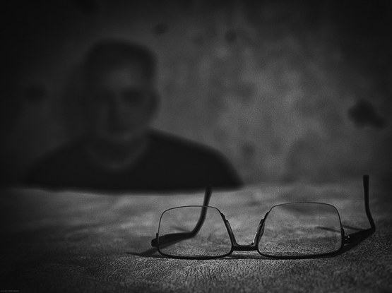 Eine Person sitzt im Hintergrund. Sie ist unschrf und man kann sie nicht genau erkennen. Im Vordergrund liegt eine Brille die sehr scharf abgebildet ist. Das Foto ist in Schwarzweiss mit einer gewissen Körnung.