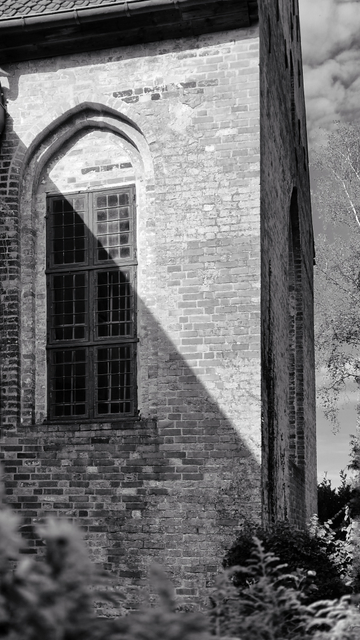 Schwarzweißfotografie eines Gebäudeteils aus gemauertem Ziegelstein. Auf der linken Seite des Fotos ist ein großes, rechteckiges Sprossenfenster zu sehen, das senkrecht eingebaut ist. Die Maueröffnung, in die das Fenster eingebaut ist, zeigt die ursprüngliche Form eines gotischen Fensters. Eine diagonale Trennlinie verläuft über die Front des Gebäudeteils von oben links nach unten rechts und teilt sie in Licht und Schatten, wobei der Schatten unten und das Licht oben ist. Rechts neben dem Gebäude sind in einem schmalen, senkrechten Streifen die Krone eines Baumes und ein leicht bewölkter Himmel zu sehen. 