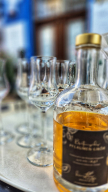 Nahaufnahme einer Flasche aus Weißglas auf der rechten Seite des Bildes sowie von Gläsern aus Weißglas, die hinter der Flasche leicht versetzt links von ihr stehen. Die Flasche ist zu etwa zwei Dritteln mit einer cognacfarbenen Flüssigkeit gefüllt. Alles, auch der Hintergrund, wirkt unscharf. 