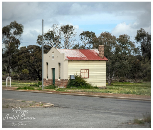 A small, historic looking building in Dumbleyung, Western Australia, with a distinctive design.

The front section is pale grey white with a small brick base and a peaked roof, while the main body is yellow cream corrugated metal with a rusty red corrugated iron roof and a brick chimney.

It sits near a paved road with a light post, surrounded by dry grass and scattered trees under a dramatic, cloudy sky.