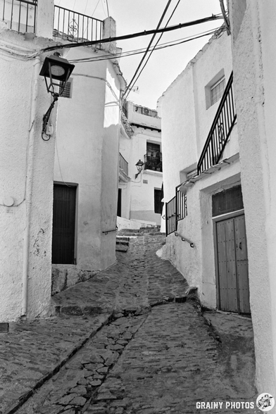 Narrow cobblestone alley with steps, lined by whitewashed buildings and balconies, overhead wires, and vintage street lamps in Capileira, a Mediterranean village, captured in black and white.