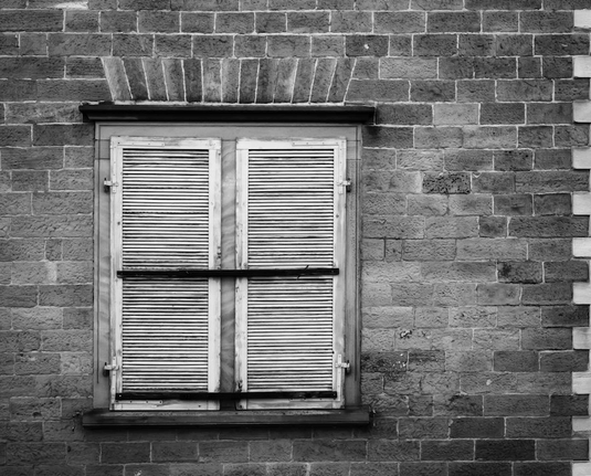 Ein altes Fenster das durch alte Fensterläden geschlossen ist. Das Ganze ist in einem Mauerwerk aus Sandsteinen zu sehen. Das Foto ist in Schwarzweiß.