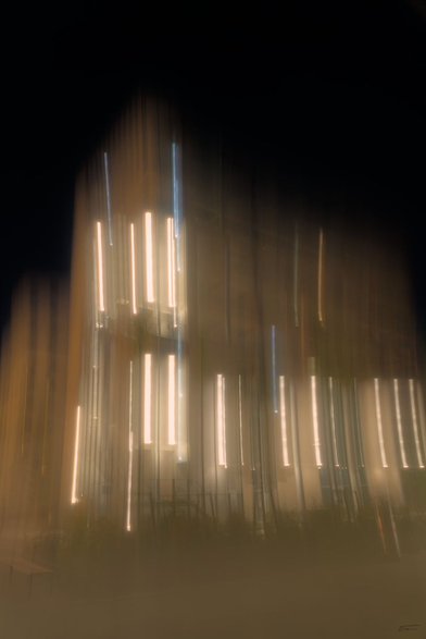 Das Foto zeigt eine nächtliche  Szene mit vertikalen Lichtstreifen, die durch Bewegung eines schlecht fixierten Statives entstanden sind. Die Lichter sind von einer Gebäudefassade, die durch die lange Belichtungszeit und die unscharfe Aufnahme zu streifigen, verschmierten Linien geworden sind. Die Farben sind warm und dominiert von Gelb- und Orangetönen der künstlichen Beleuchtung. Der Hintergrund ist dunkel, was den Kontrast zu den Lichtern verstärkt.
Die Unschärfe und die Lichtspuren verleihen dem Bild einen dynamischen, fast abstrakten Charakter. Es wirkt fast wie eine künstlerische Darstellung von Licht und Bewegung.