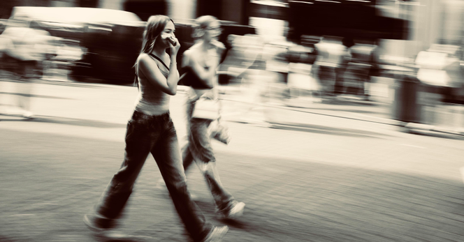 Slightly blurry moving camera photo of two young girls in jeans striding down the street, one holding a phone to her ear. 
