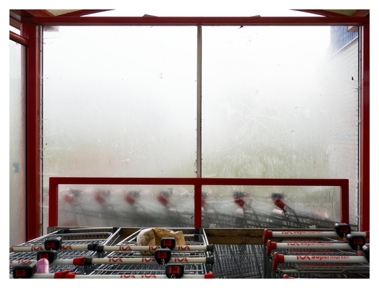 En liggande bild på ett fönster på en plats för vagnar vid en ICA-affär. Det är disigt och vagnarna på andra sidan fönstret är oskarpa pga diset och fönstret. Röda balkar ramar in vagnarna, en papperspåse och en rosa burk ligger på ett par av vagnarna.