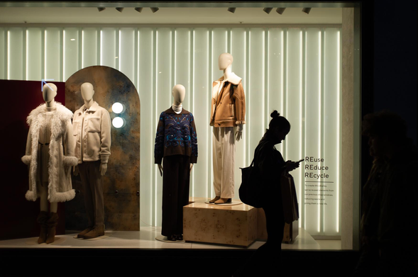 The image captures a shop window display at night. The window is brightly lit, showcasing mannequins dressed in various outfits. The background of the display features vertical, light-emitting panels that create a clean, modern aesthetic.

Three mannequins are positioned in the window. The first, on the left, is wearing a long, cream-colored coat with a fur trim, over a dress, and brown boots. The second mannequin is dressed in a light-colored jacket and trousers. The third mannequin, slightly to the right, wears a blue patterned sweater with dark pants. The final mannequin wears a brown jacket and white trousers.

A woman, silhouetted against the bright window, stands to the right of the display, looking at her phone. Her dark hair is pulled up, and she carries a large bag. Another person is also silhouetted in the right side of the image, observing the display.
