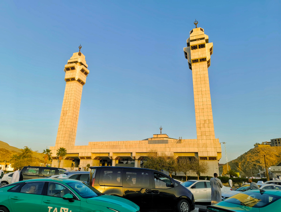 The Masjid-e-Ayesha (Ayesha's Mosque) in Makkah (Mecca) Saudi Arabia, a modern-looking mosque with a rectangular main building and two prominent, rectangular minarets with multiple levels. The sky above is clear blue, and several cars, including green taxis and a black van, are parked in front of the mosque. A person in a white thobe walks near the cars.