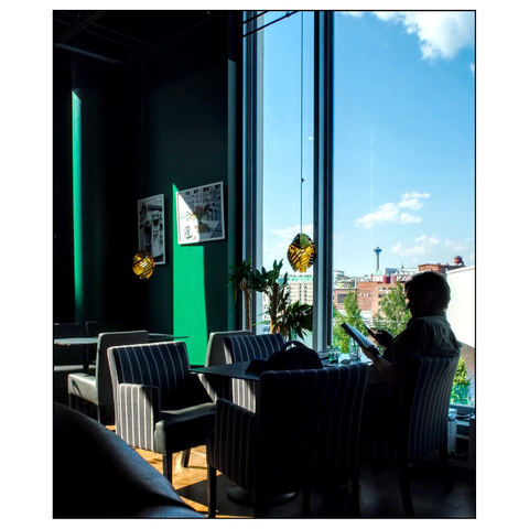 The image shows the interior of a modern café lounge. The space features large floor-to-ceiling windows that offer a panoramic view of an urban landscape, including buildings and a prominent tower in the distance. The sky outside is clear with scattered clouds.
Inside, the seating area includes several upholstered chairs with striped patterns, arranged around small tables. A person is seated near the window, facing the view and holding a book. The walls of the café are painted in green tones, with one section highlighted by the sun. Decorative elements include framed pictures and hanging light fixtures with a geometric design. There are also potted plants placed near the windows. (draft by mistral, edited by author)