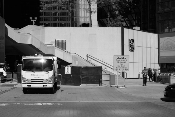 Looking across the street at the base of an office tower. At left, we see the front end of a parked truck with a bright flare of sunlight on its windshield. in the background is a long, angular modern staircase leading to the upper left. Right of centre is a sign announcing a street closure. At far right are a couple of people, small in the scale of the photo, standing on the pavement.
