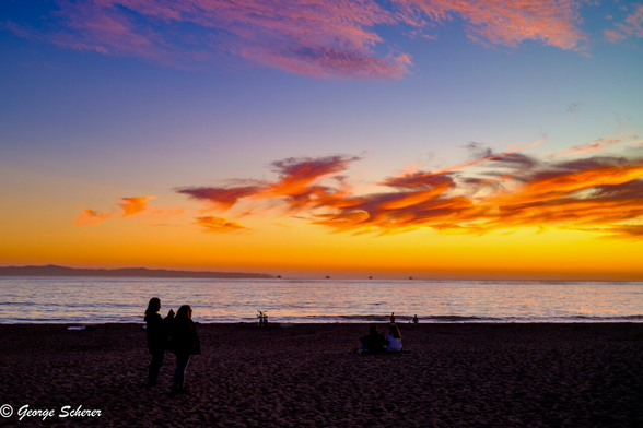 View of the sunset looking across a dark, sandy beach.  The sky is orange near the horizon, shading into blue near the top of the image.  The clouds are lit with bright orange from underneath.  There are a couple of people in the foreground, seen in silhouette, taking photos of the sunset with their phones.