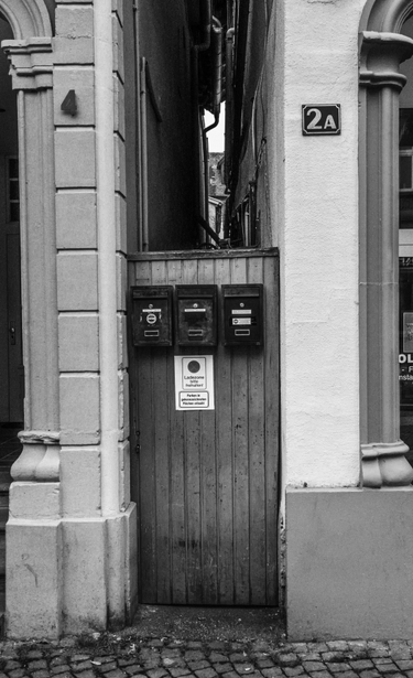 Man sieht eine kliene schmale holztür, die zwischen zwei Geschäften liegt. Drei Breifkästen sind dort angebracht. Man kann oberhalb der Tür sehen das dahinter eine sehr schmale Gasse liegen muss. Das Foto ist in Schwarzweiß.
