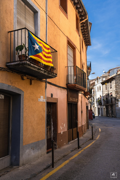Color photo of a curving street in the Catalan town of Besalú. On a balcony railing in the left foreground is hanging the Catalonian Estelada flag with horizontal red and yellow stripes and a blue triangle with a single white star on the left side.