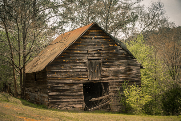Weathered two-story wooden barn with dark, aged horizontal plank siding and a deteriorating orange-brown metal roof. The structure features a small square opening or window on the upper level and a darker rectangular doorway or opening at ground level. Dry grass and scattered fallen leaves cover the ground in front of the barn. The building is surrounded by bare and leafing deciduous trees under an overcast sky, with dense woodland visible in the background. The barn shows significant age and decay, with warped siding and a sagging roofline.
