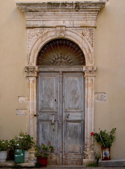 An ornate, weathered wooden door with intricate detailing and an arched top is surrounded by decorative pillars. The door features a rustic, faded blue hue. Potted plants in colorful containers are placed at the base, adding a touch of greenery.