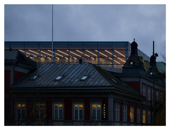 En liggande bild på ett gammalt trähus med tända lampor i taket synligt genom fönstren. Bakom den ett nybyggt, kantigt hus med panoramafönster och lysrör i taket. En molnig himmel i skymningen.