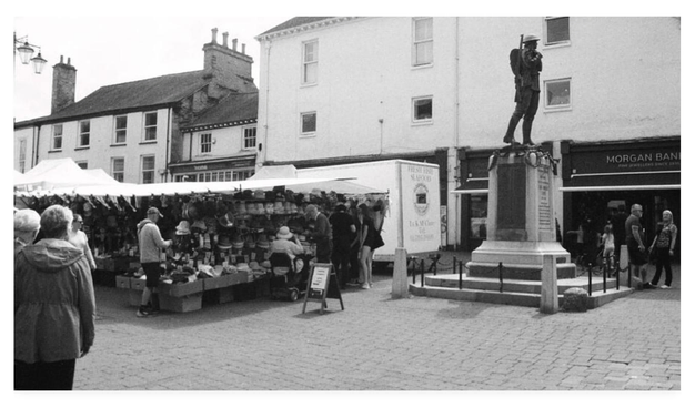 Black and white film photograph of a bustling market square and a stall selling headgear.  To the right of the shot is a war memorial statue.