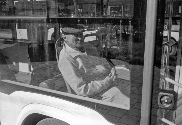 The black-and-white photograph shows an older man sitting inside a bus, seen through the window. He wears a flat cap and a light-colored jacket, his hands resting together in his lap. The glass reflects the street outside — pavement tiles, parts of another bus, and some buildings — blending interior and exterior scenes. The image captures a quiet, contemplative moment, emphasizing texture, light, and reflection in a classic documentary style.
