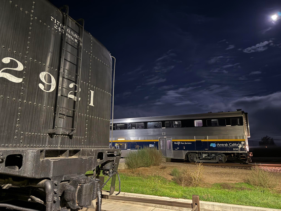 A tale of two trains. In the foreground is the back of the Modesto Bee steam locomotive with the number 2921 painted in white. In the background is the front of a modern Amtrak trail. Cloudy skies are illuminated in the darkness behind.