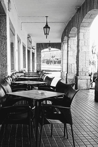 Leere schwarze Cafétische und Stühle in einer Galeria in Spanien in Zentralperspektive. Rechts die Bögen aus Granit, links Fenster in der Hauswand mit Rahmen aus Granit. Der Boden ist dunkel gefliest mit kleinen Fliesen. An der Decke hängt eine altmodische, schwarze, eiserne Lampe