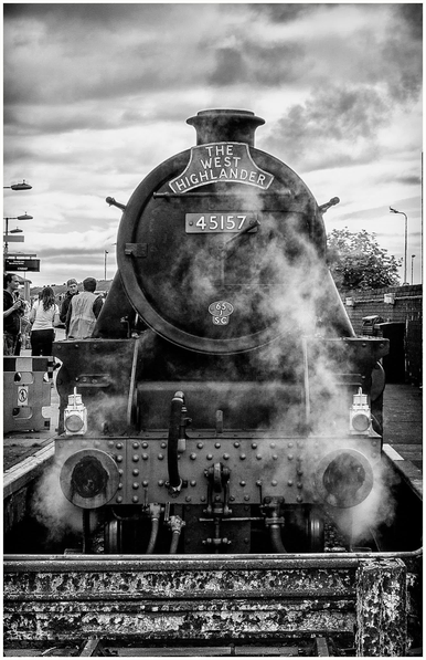 A vintage steam locomotive labeled "THE WEST HIGHLANDER" with the number "45157," emitting steam. In the background, people are seen on a train platform, with a cloudy sky above.