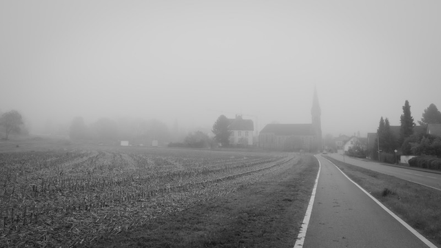 Von Rechts unten mommt eine Strasse und ein Fahrradweg. Bei führen in einen Ort. Links daneben ich ein Ackerfeld zu sehen. Vom Ort sieht man wegen Nebel kaum was nur eine Kirche im Dunst ragt auf. Das Foto ist in Schwarzweis im 16:9 Format.