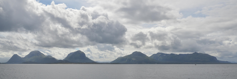 A panoramic photo of water leading to mountains. The sky is filled with clouds.