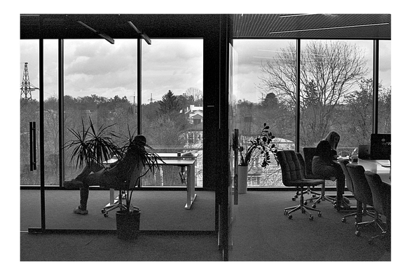 A grainy, black and white film photograph of an office interior with large windows. The image is split by a wall: on the left, a person relaxes with feet on a desk, looking out; on the right, another person works at a desk.