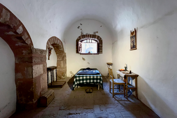 The image depicts the interior of a simple, possibly historical, room. The room has a vaulted ceiling and appears to be constructed of stone and stucco. The walls are white, with some areas showing signs of age and wear, such as peeling paint. The floor is made of reddish-brown stone tiles.

The room is sparsely furnished. The central element is a bed covered with a green and white plaid blanket. There is an open window above the bed.  A small table with a candle and some fruit sits near the window, along with a wooden chair.  On the opposite side, an archway leads to another area. Underneath the archway, there's a small wooden table, a wooden cross, and a small chest.  A painting is hanging on the wall. The room's minimalist decor and religious imagery suggest it might be a monastic cell or a similar type of dwelling.

The lighting appears to be natural, coming from the window, casting shadows on the floor. The overall atmosphere is one of simplicity, quietude, and perhaps a sense of history.