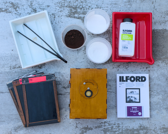 A flat lay of my pinhole camera, film holders, developing tanks, chemicals, and photo paper.