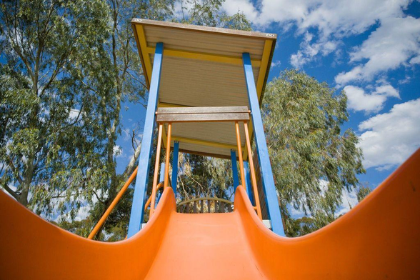 Wide-angle photo looking up from the bottom of a bright orange coloured children's slide in a local park.