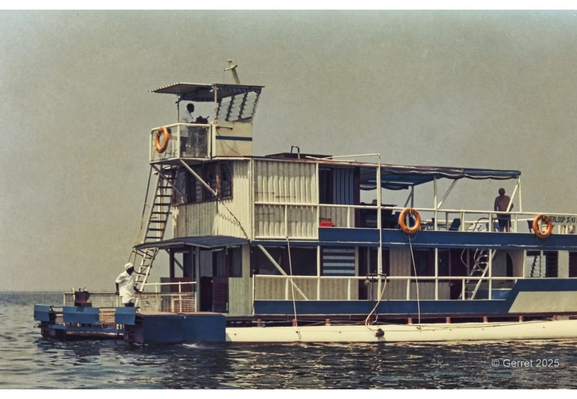 A vintage houseboat floats on calm water under a hazy sky. The structure is multi-leveled with lifebuoys and a man standing on the deck, conveying tranquility.