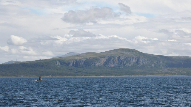 A photo of water leading to the cliff side of a mountain. There's a small tower in the water to the left of the photo. The sky is filled with clouds.