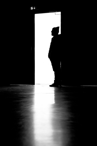 Backlit shot. A man stands in the doorway of a dark room and looks into it. His silhouette is perfectly outlined in profile, as bright sunlight shines through the door from outside. The shiny floor gently reflects the silhouette.