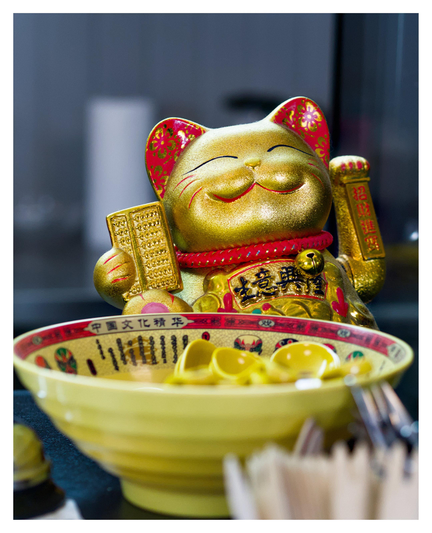 En stående bild på en leende och vinkande katt i guld och rött på en sushi-restaurang. I förgrunden en gul och röd skål med gula saker. Det övriga är ur fokus.