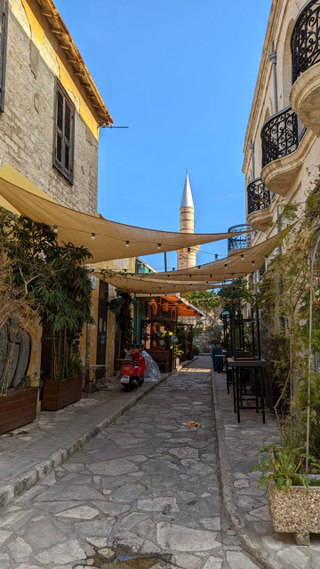 Das Bild zeigt einen Blick in eine enge mediterrane Gasse. Die mit viel Grün geschmückte Straße wird von dreieckigen Sonnensegeln überspannt. Im Hintergrund ist ein Minarett vor blauem Himmel zu erkennen.