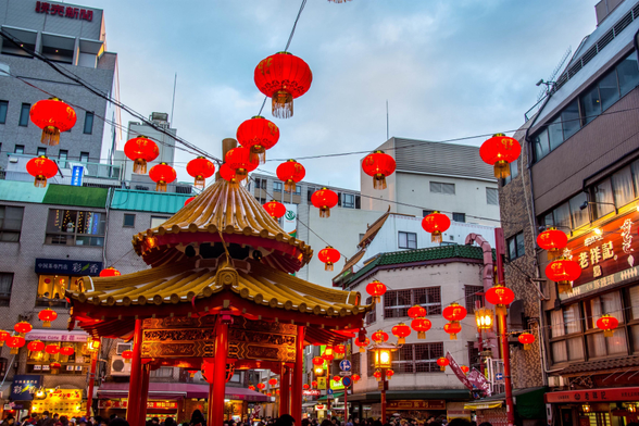 A twilight view of Kobe Chinatown in Japan. Dozens of red lanterns hang above a golden pavilion surrounded by buildings with signs in Chinese and Japanese. People walk below, illuminated by the warm glow of evening lights.