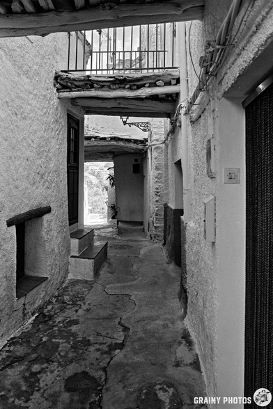 A narrow, old stone alleyway with textured white walls and wooden tinao overhead—typical of Capileira—leads to a sunlit area. A balcony with railings is visible on the tinao, whilst doors and small windows line the narrow passage.