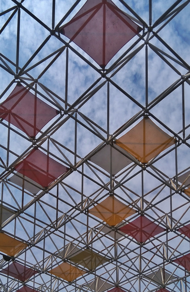 Geometric metal grid ceiling with colorful diamond panels in red and orange against a cloudy blue sky. (colored)