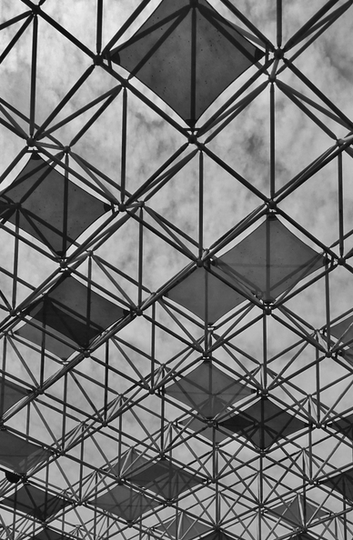 Geometric metal grid ceiling with colorful diamond panels in red and orange against a cloudy blue sky. (black and white)